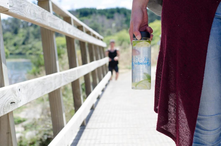 cropped shot of woman in red cardigan holding water bottle with lime slices while walking over wooden bridge with runner in background