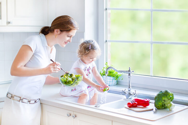 mother-holding-bowl-of-salad-while-watching-toddler-girl-sit-on-counter-and-wash-lettuce-leaves