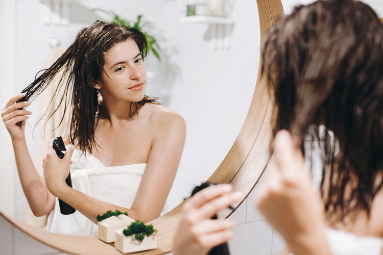 woman-spraying-hair-with-spray-bottle-while-standing-in-front-of-mirror-wearing-towel