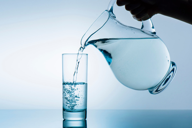 A hand pouring water from a glass jug into a tall glass with a pale blue grey background and surface.